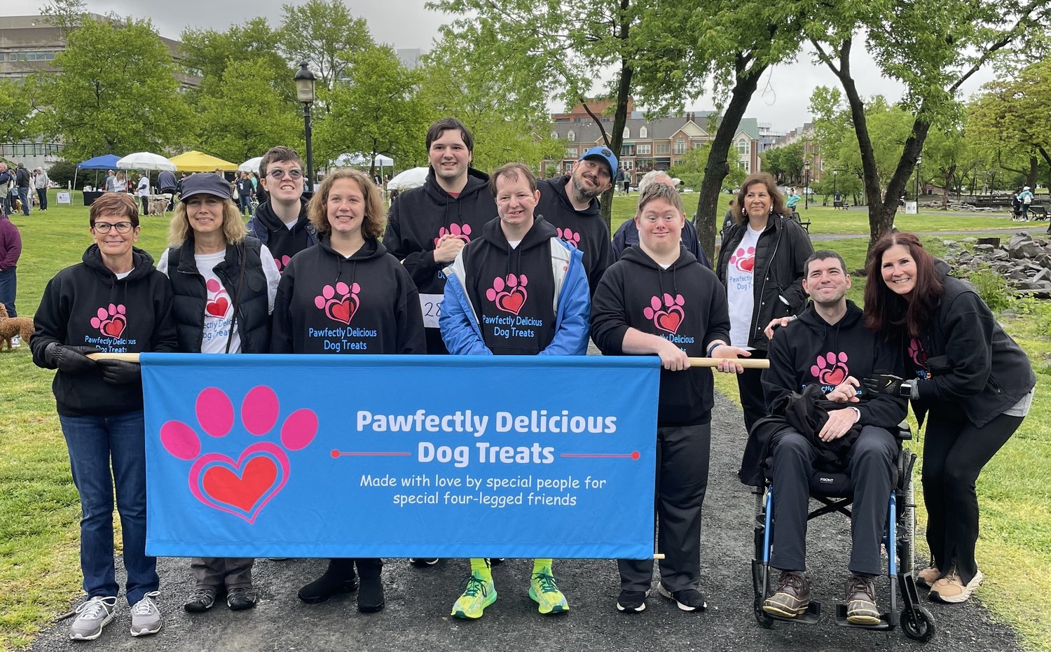 A group of Pawfectly Delicious Dog Treats team members, who are a nonprofit partner of Dr. Phytos, smiling and holding a banner at an outdoor community event promoting inclusive employment for adults with disabilities and handmade natural dog treats.